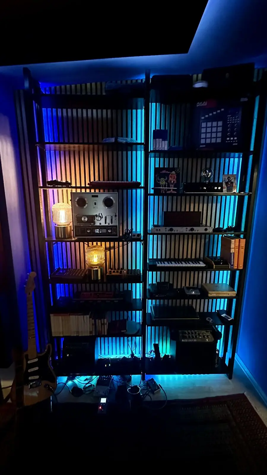 Floor-to-ceiling shelf against the slatted wall, lit blue and warm: tape machine, drum machine, preamps and keyboards, a guitar leaning at the left