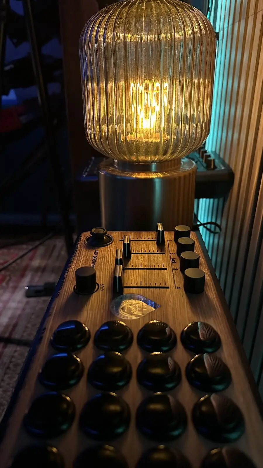 A wooden-cased sampler in the foreground, sixteen pads, faders and knobs, a warm ribbed lamp above it